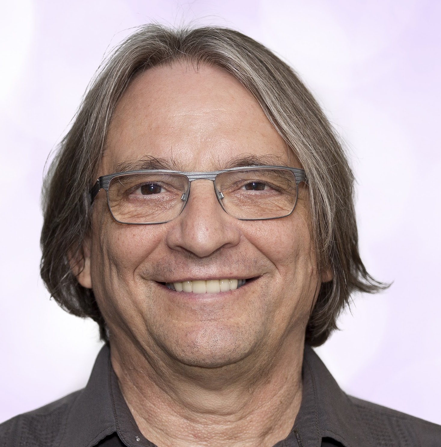 A middle-aged man with shoulder-length gray hair and glasses smiles at the camera. He is wearing a dark shirt and is posed in front of a light, softly blurred background.