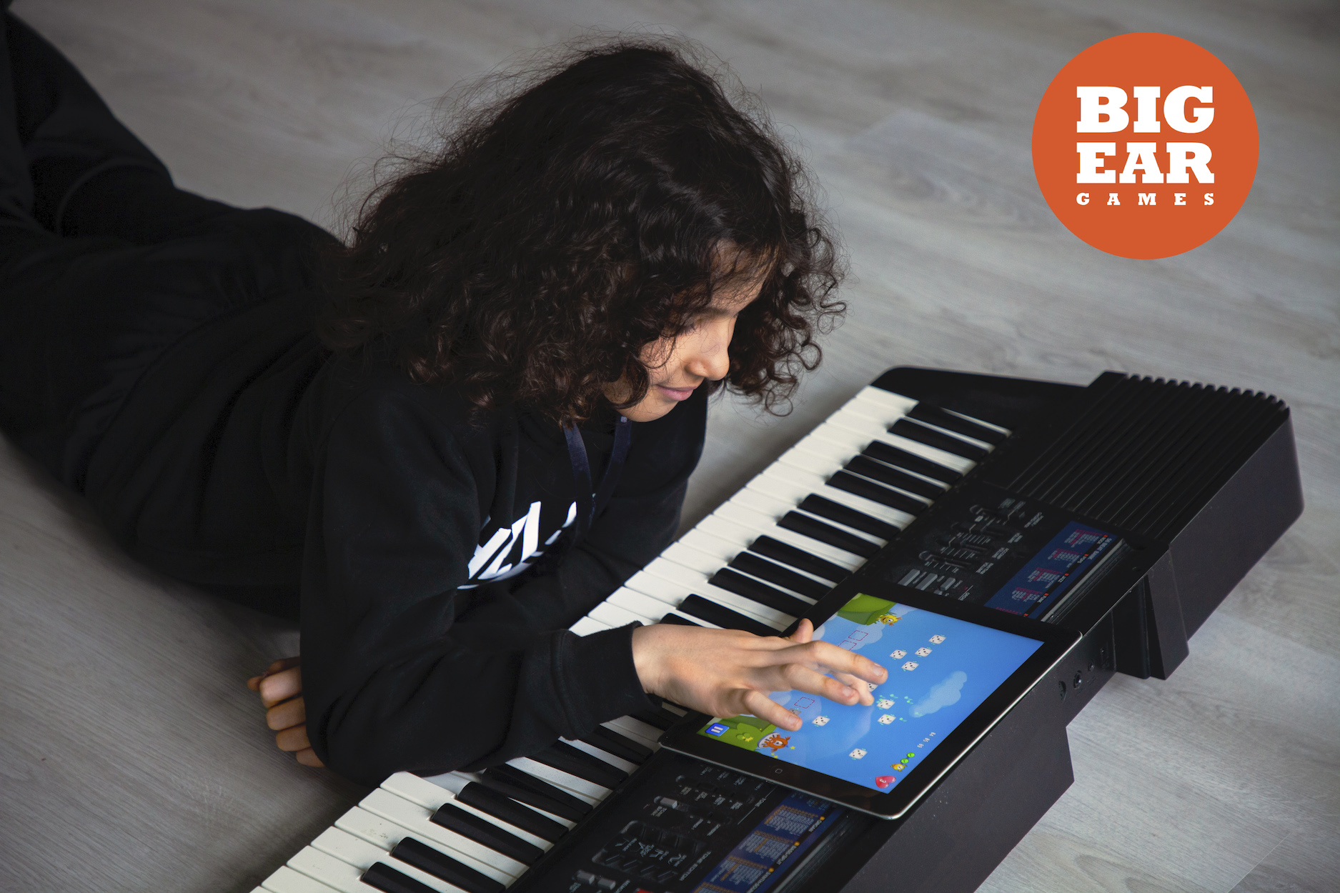 A child with curly hair lies on the floor using a tablet screen embedded in a keyboard. In the top right corner, there is an orange BIG EAR GAMES logo.