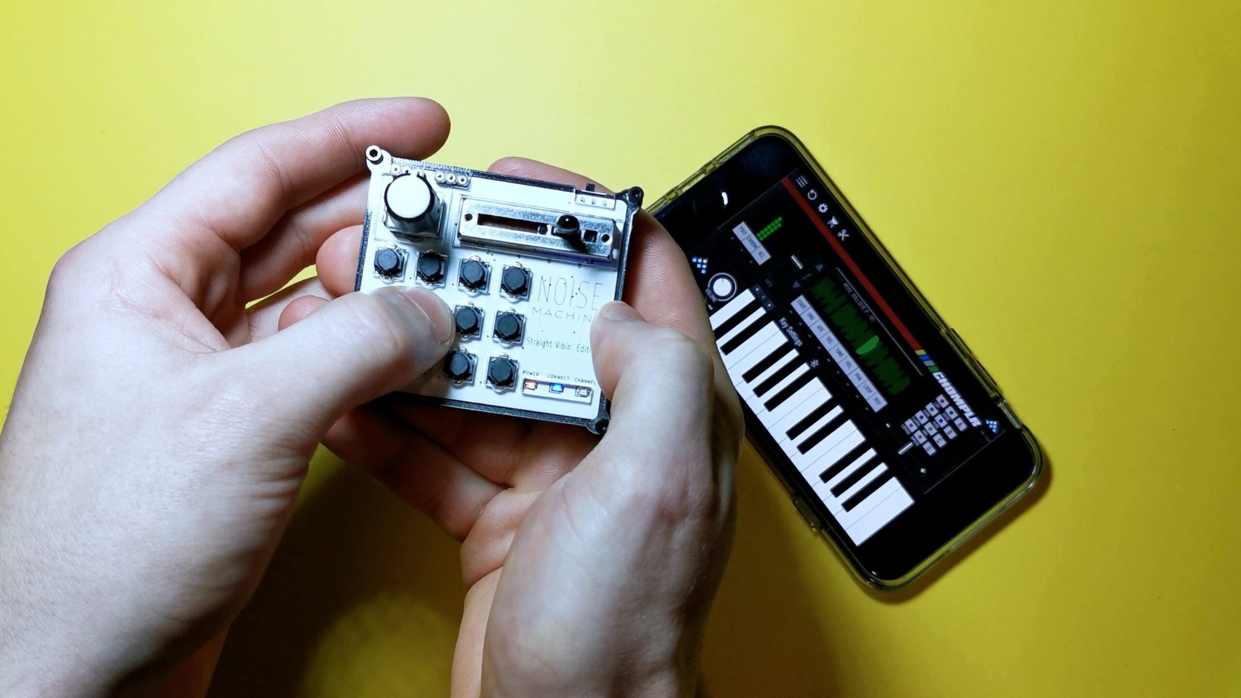 Two hands holding a small music synthesizer with knobs and buttons, next to a smartphone displaying a piano keyboard app, all on a yellow background.