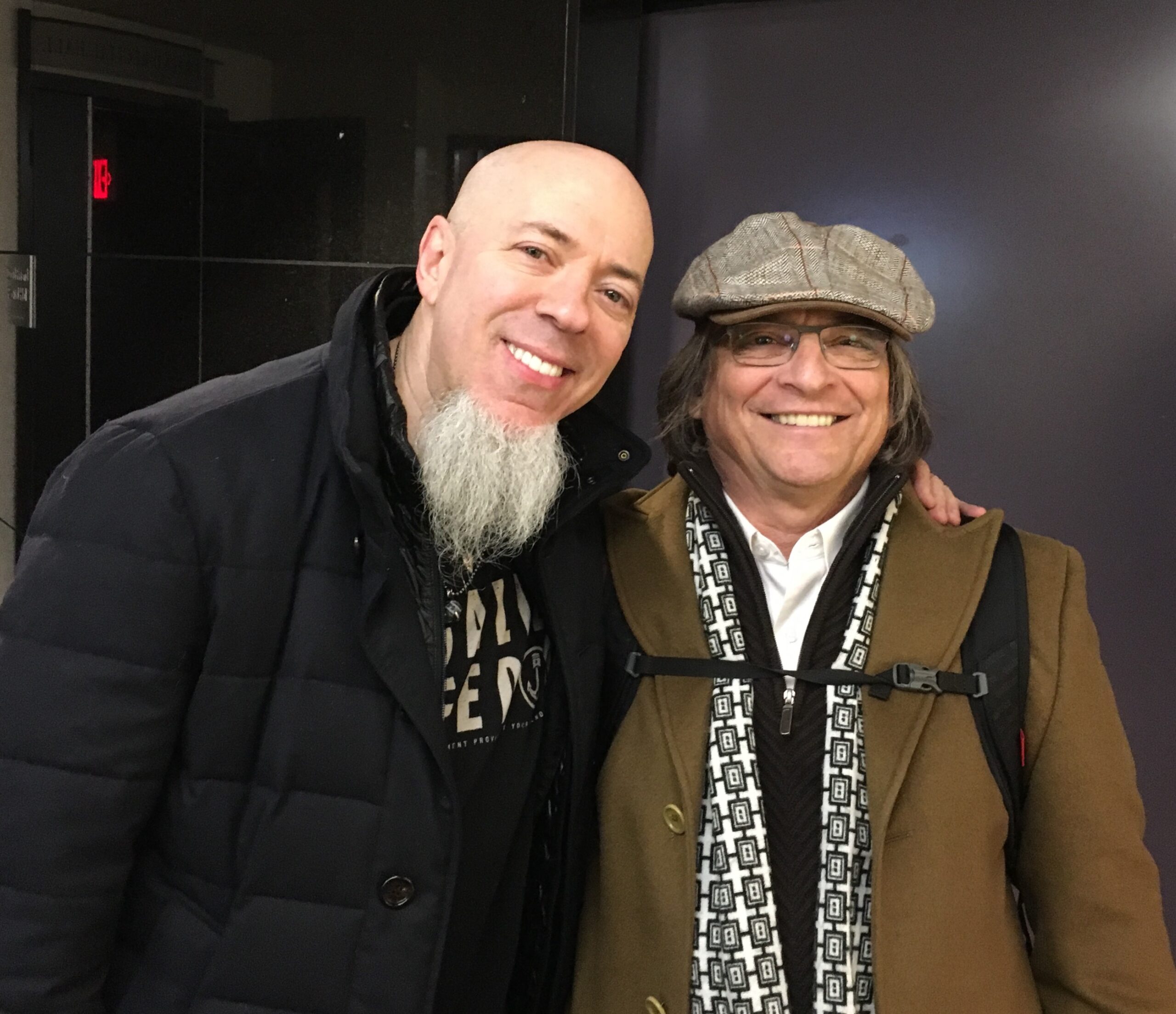 Two smiling men stand together indoors. One is bald with a long white beard, wearing a black coat. The other has glasses, long hair, a flat cap, and a brown coat with a patterned scarf and backpack.
