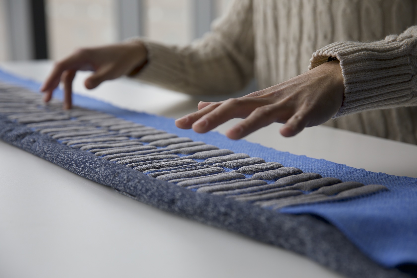 A person wearing a beige knitted sweater hovers their hands over a textured blue and gray textile laid out on a white surface, possibly feeling or interacting with its raised elements.