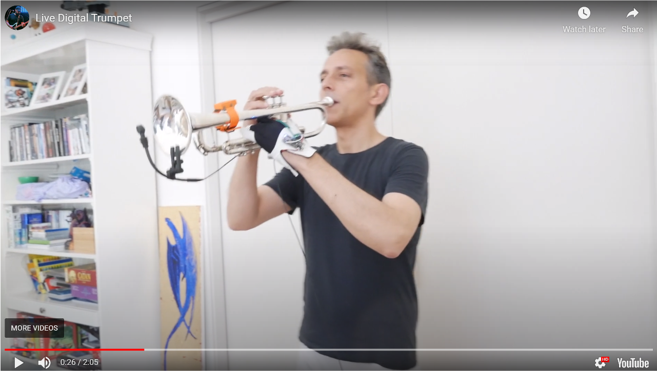 A person in a black t-shirt plays a silver trumpet indoors. Shelves with books and boxes are on the left, and a blue abstract painting leans against the wall in the background.