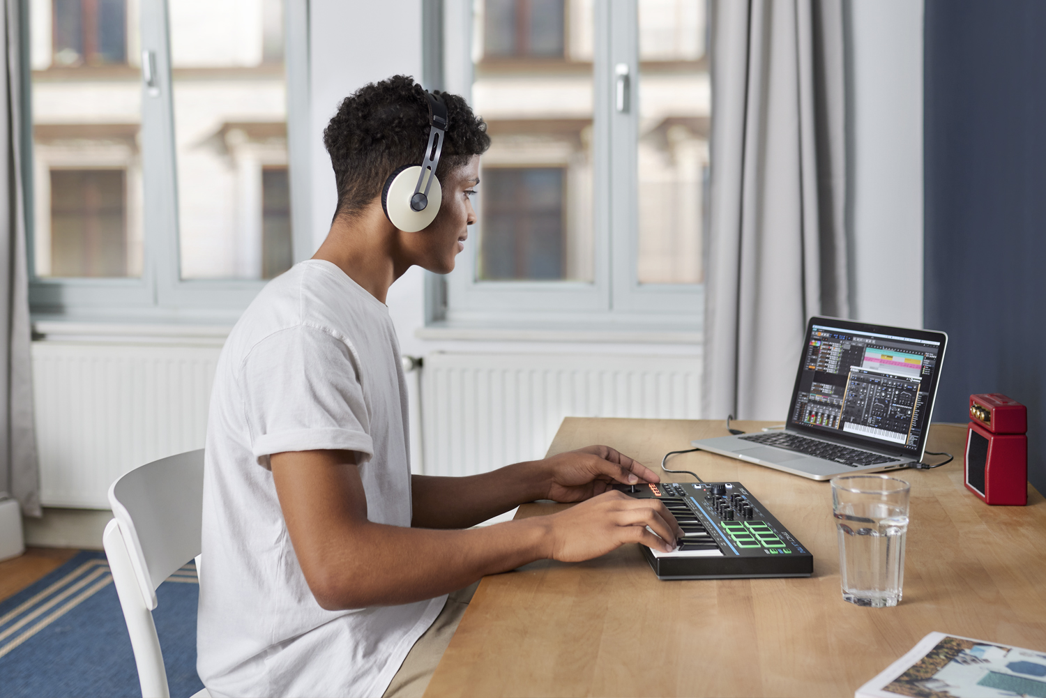 A person wearing headphones sits at a desk using a MIDI keyboard connected to a laptop with music production software. There is a glass of water and a small red speaker on the desk. Large windows are in the background.