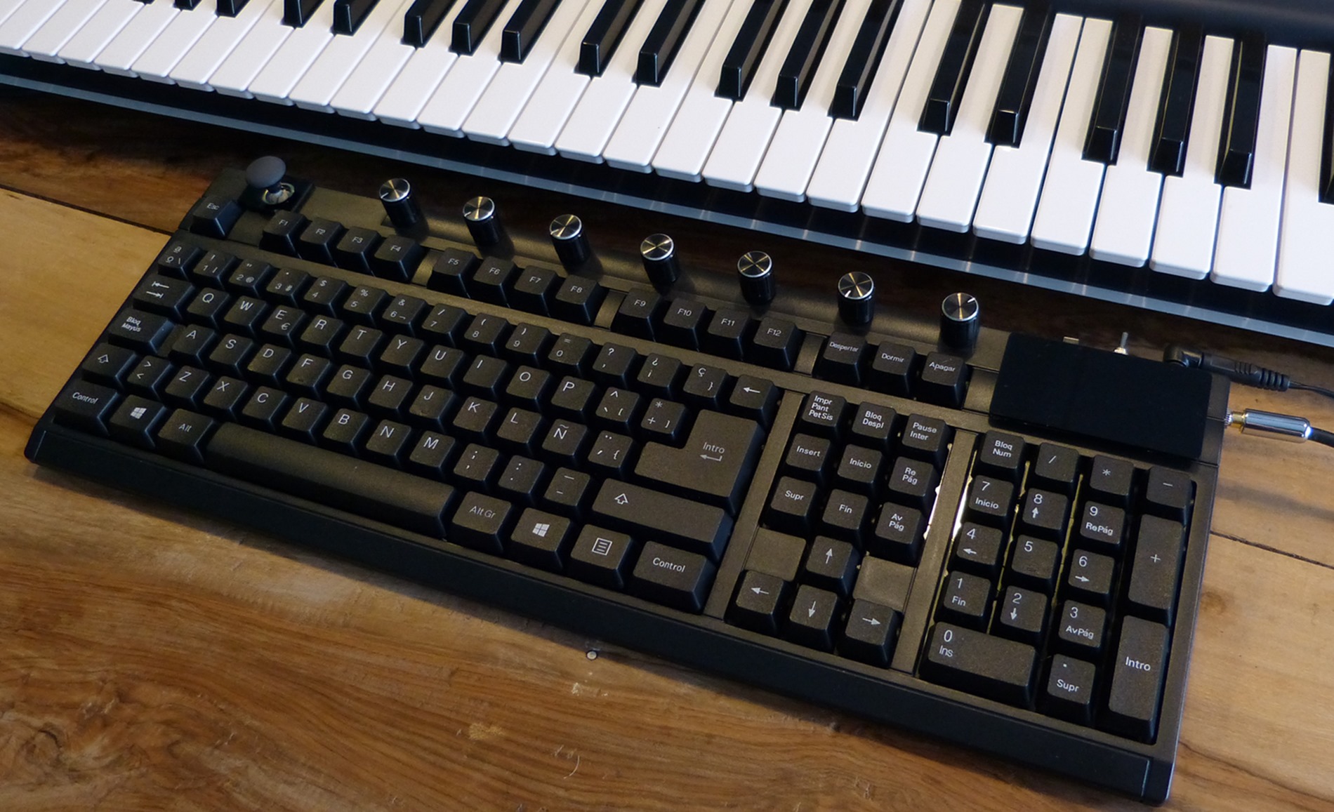 A computer keyboard with multiple dials sits on a wooden desk below an electronic musical keyboard.