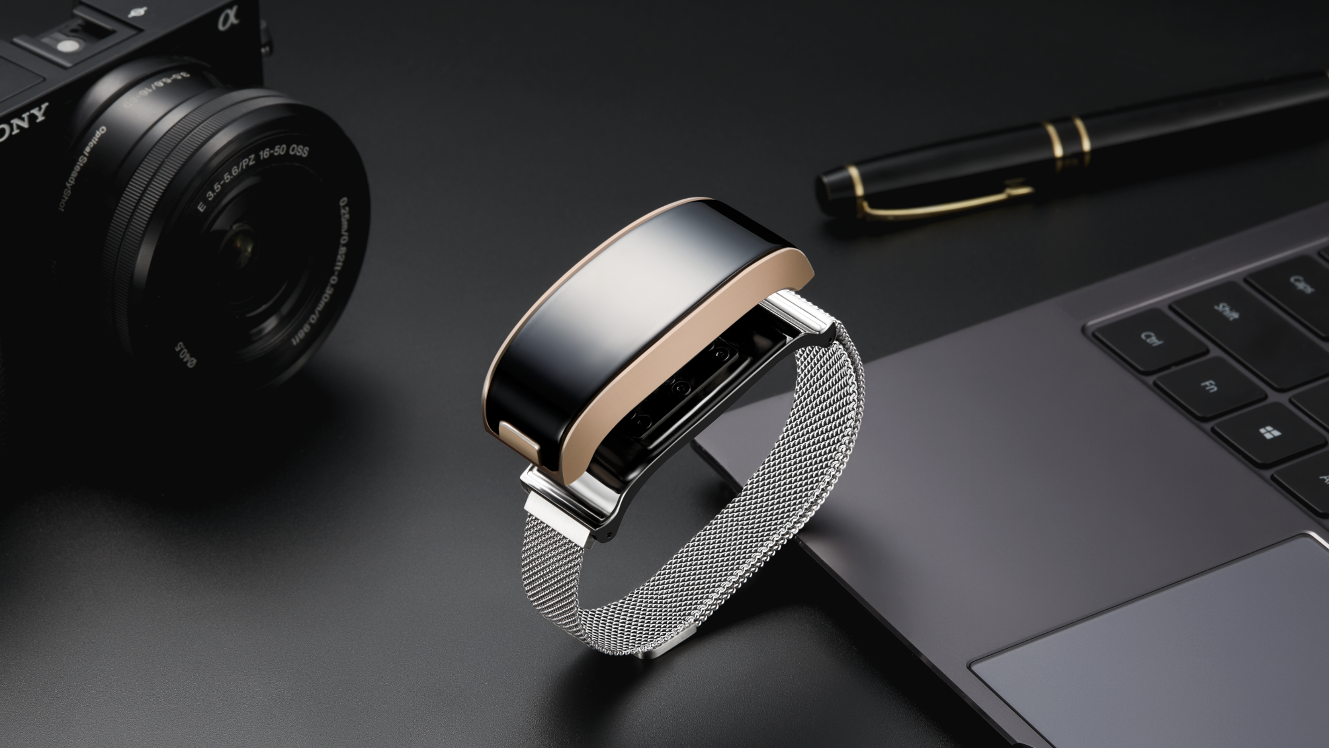A stylish smartwatch with a metallic mesh band rests on the edge of a laptop keyboard, with a black pen and a camera positioned nearby on a dark surface.