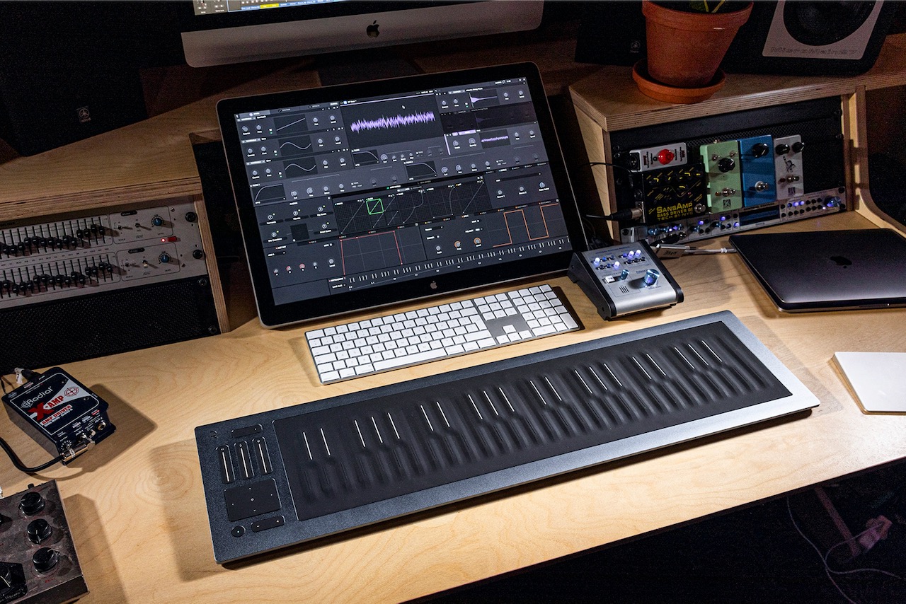 A music production desk with a touchscreen monitor showing audio software, a keyboard, a touch-sensitive MIDI controller, various audio equipment, and guitar pedals, set on a wooden surface.