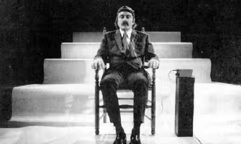 A man in a suit and tie sits in a wooden chair on a stage with tiered steps behind him. He wears a headband and is connected to a device by wires. The setting appears formal and theatrical.