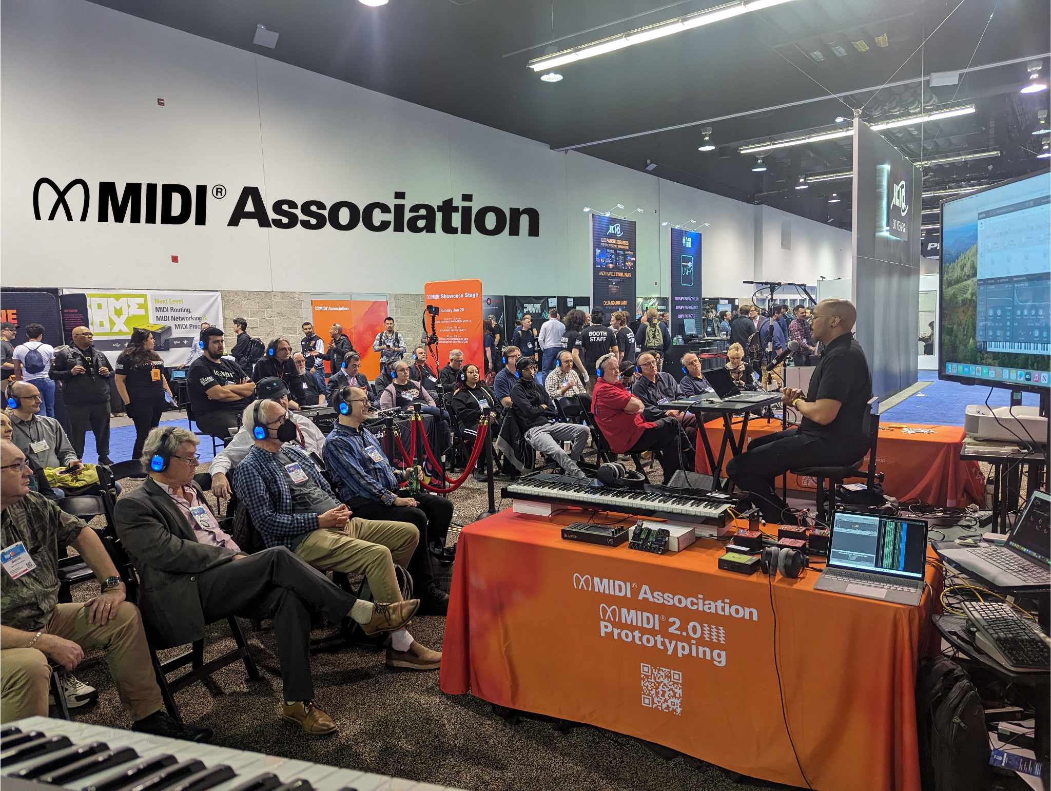 A presenter demonstrates music technology to an audience wearing headphones at a MIDI Association event. Attendees sit and watch near booths and screens in a busy convention hall.