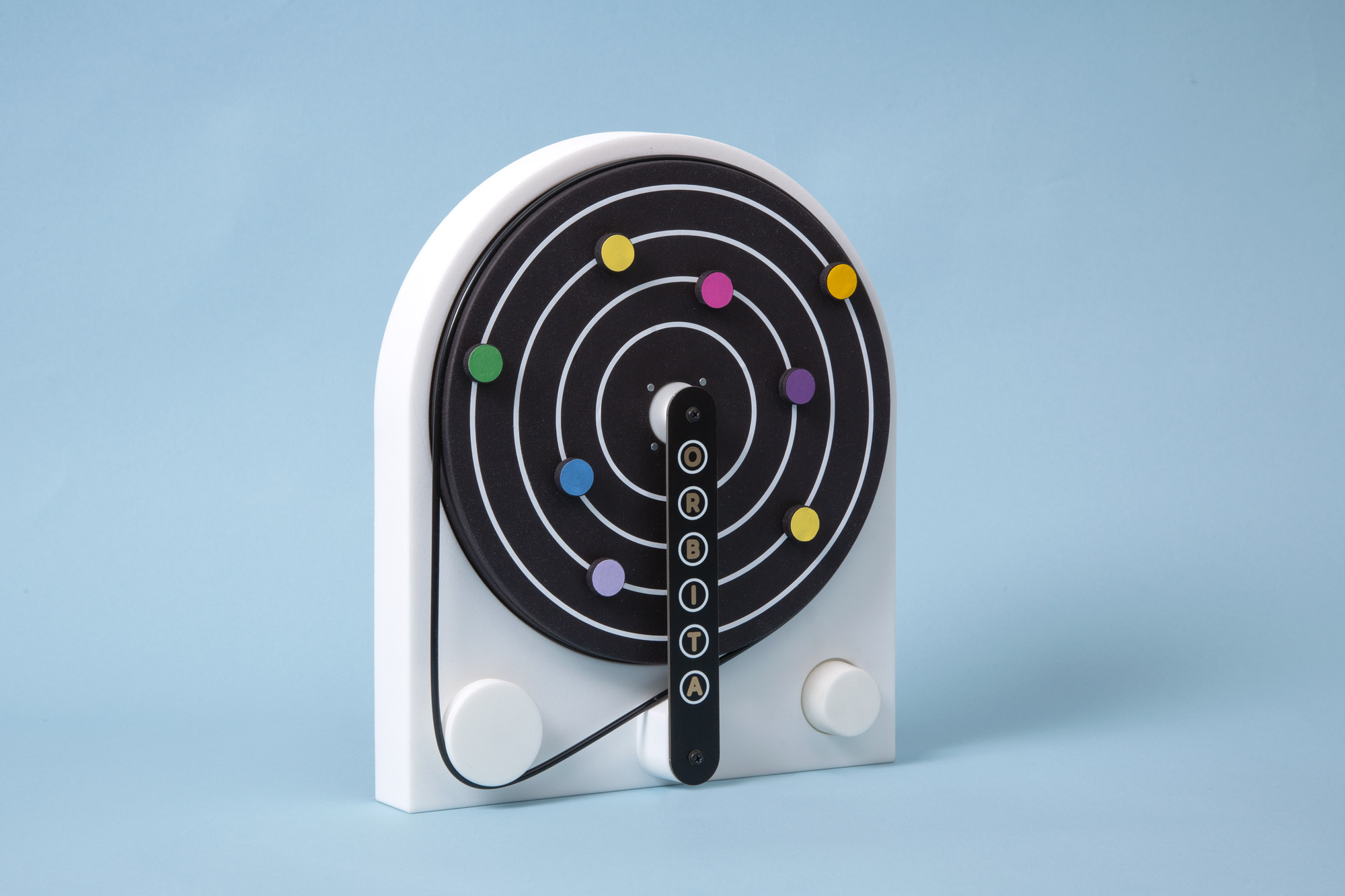 A white and black circular target with concentric rings and colorful pins is mounted on a white stand against a light blue background. The stand has two knobs at the bottom.