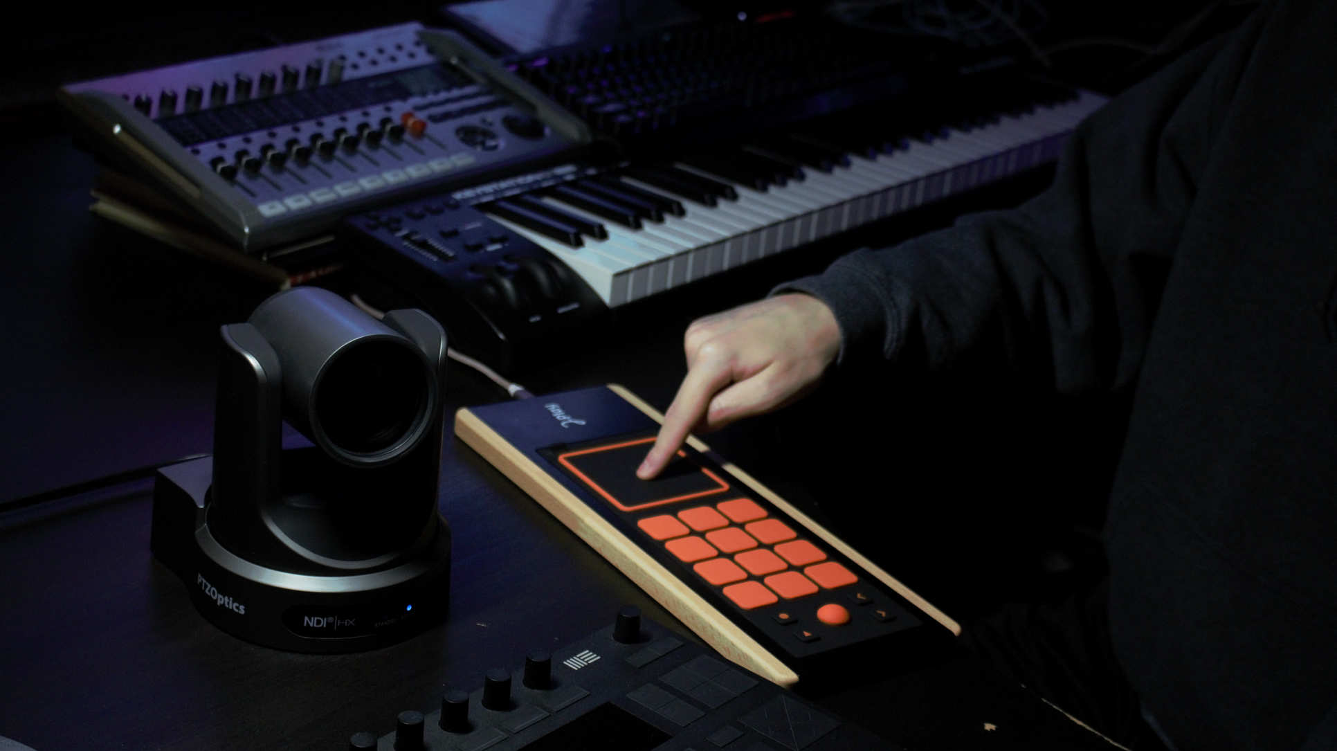 A person uses a touch-sensitive MIDI controller with orange pads, next to a digital camera and a keyboard synthesizer, in a dimly lit music studio.