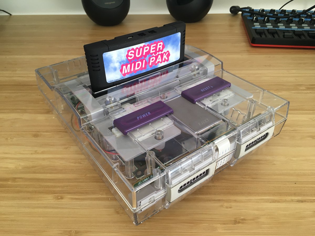 A transparent Super Nintendo console with purple buttons and a cartridge labeled Super MIDI Pak inserted, placed on a wooden surface with audio equipment in the background.