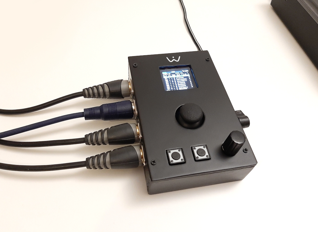 A black electronic device with a small display screen, two buttons, a knob, and four connected cables sits on a white surface. The screen shows a list of settings or options.