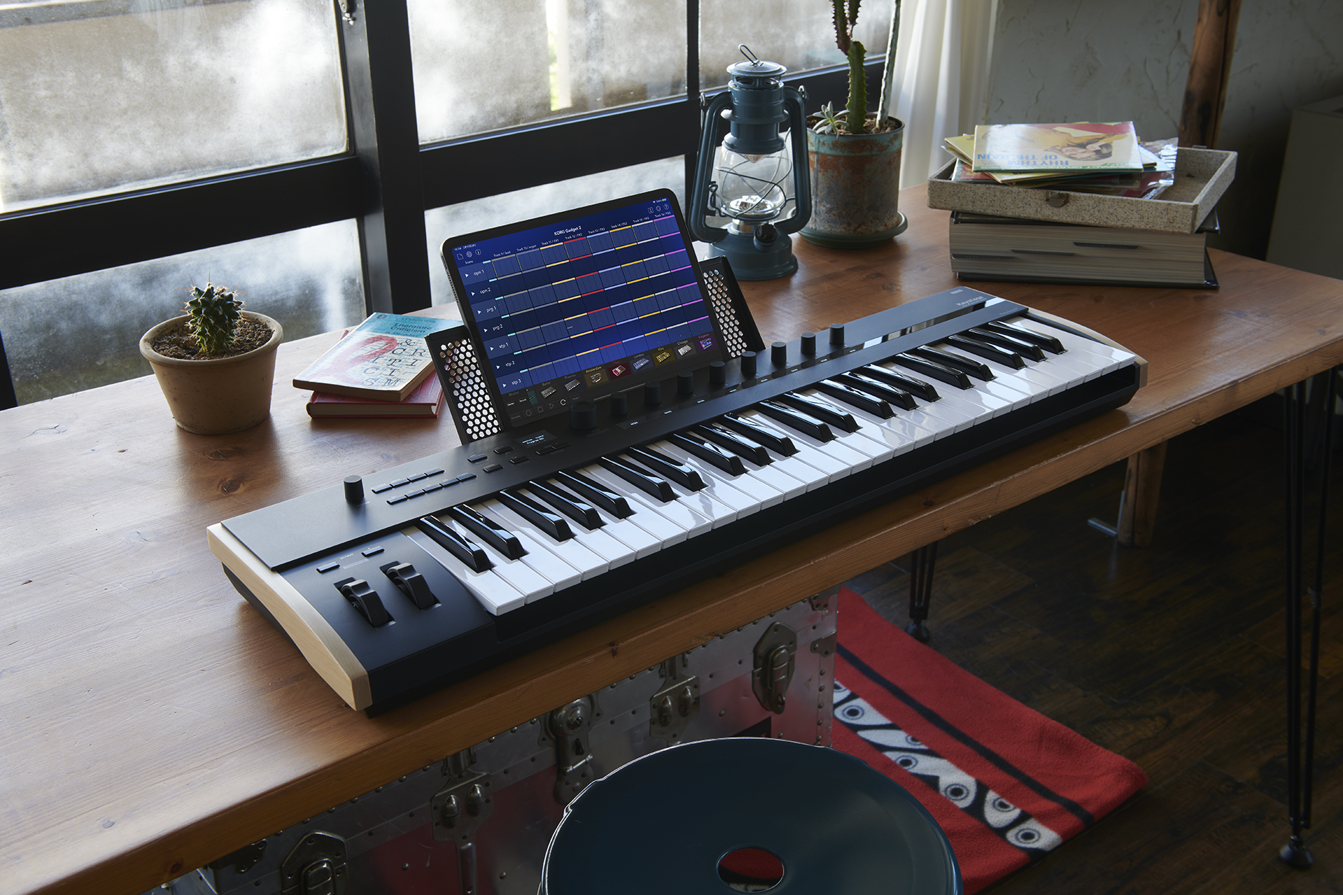 A digital keyboard with an iPad displaying music software sits on a wooden desk near a window, surrounded by potted plants, books, and a blue lamp in a cozy room.