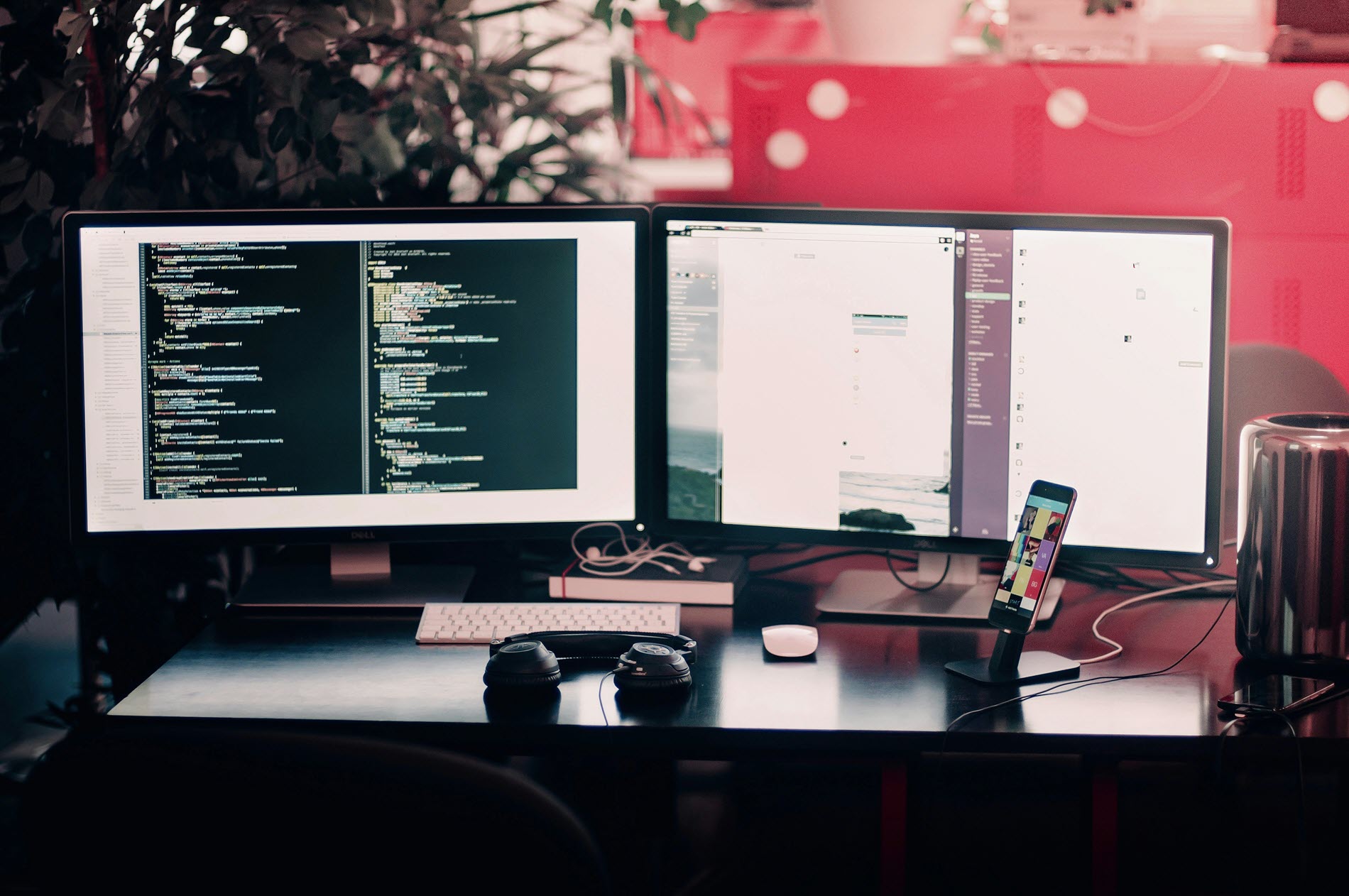 A workspace with two computer monitors displaying code and applications, a keyboard, mouse, headphones, a smartphone on a stand, and a cylindrical computer on a black desk with plants and red decor in the background.