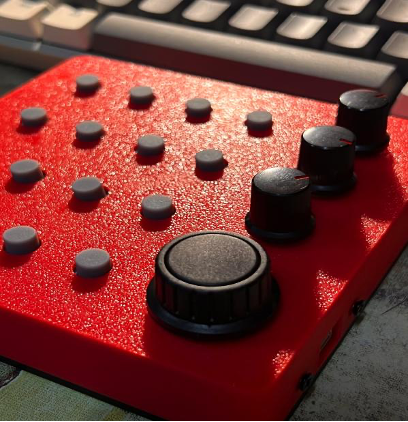 A close-up of a red audio or MIDI controller with several gray buttons and black knobs, positioned on a desk with part of a keyboard visible in the background.