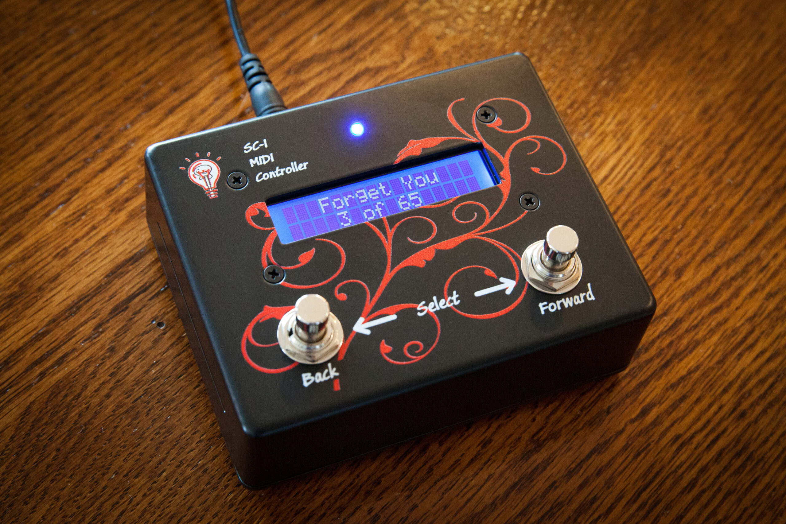 A black MIDI controller with a blue screen displaying Forget You sits on a wooden surface. It has two footswitches labeled Back and Forward, a Select button, decorative red designs, and an indicator light.