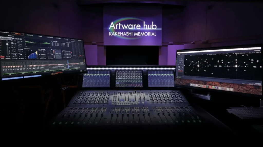 A dark, modern audio control room with two large computer screens and a mixing console. The back wall features illuminated text: Artware hub KAKEHASHI MEMORIAL in white and purple lighting.