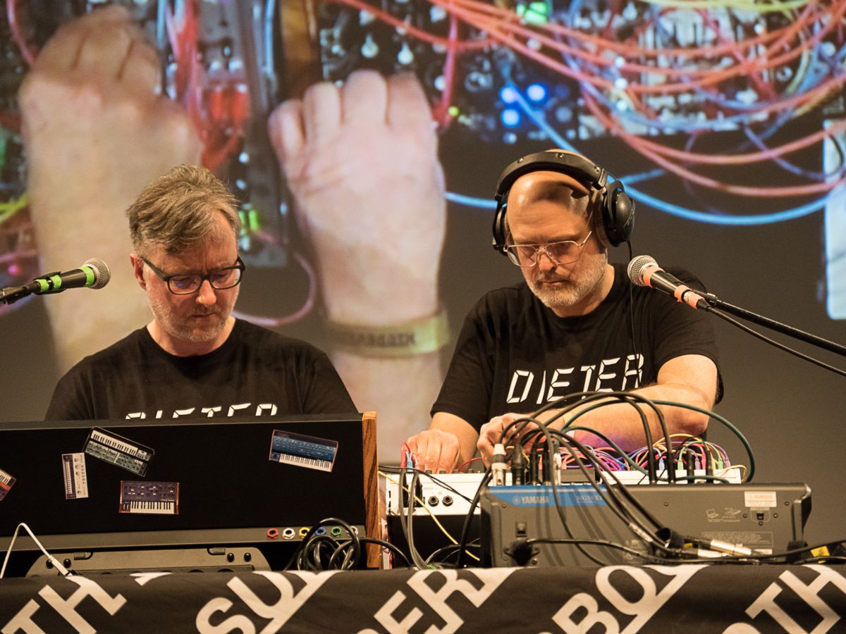 Two men wearing DIETER shirts perform electronic music on stage, using synthesizers and cables, with a large projected image of their hands working on equipment in the background.