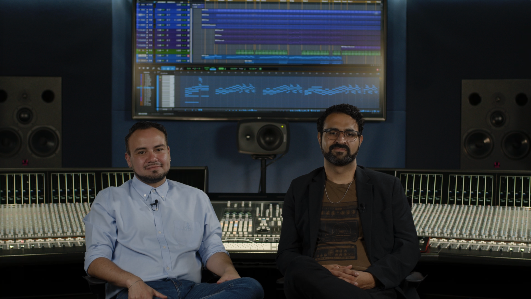 Two men sit in a recording studio in front of a large mixing console and speakers, with a computer screen displaying music production software in the background.