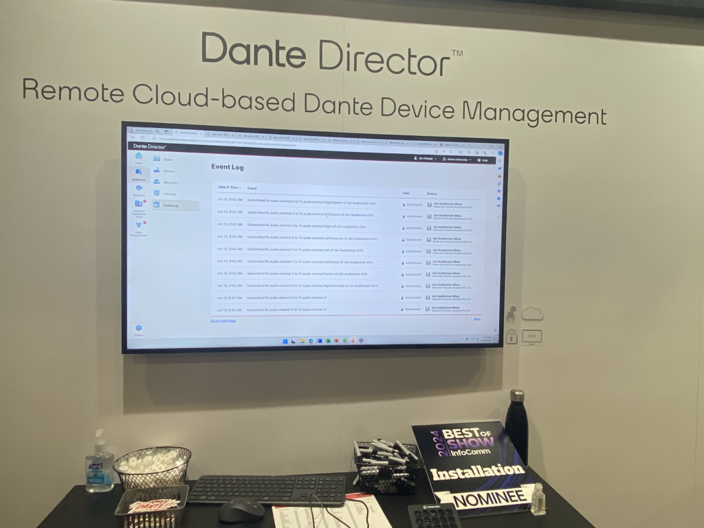 A monitor displays the Dante Director event log screen on a wall labeled Remote Cloud-based Dante Device Management. Below are a keyboard, sanitizer, brochures, pens, a water bottle, and an award sign.
