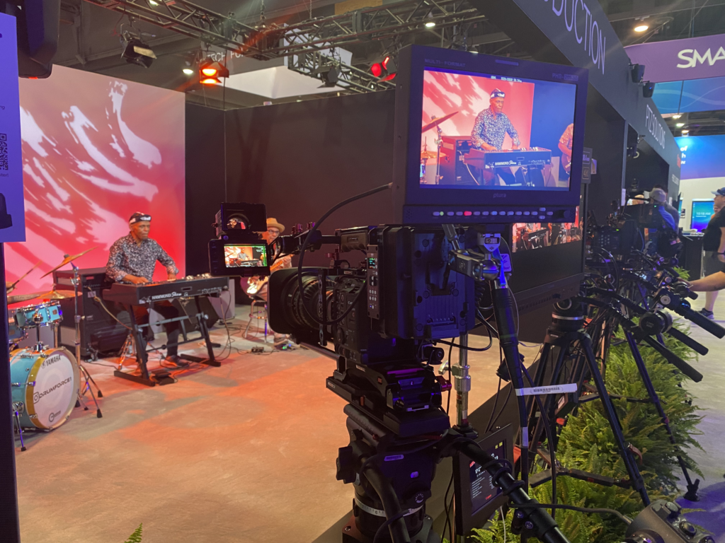 A stage with a musician playing a keyboard is filmed by several cameras. The main camera’s monitor shows a close-up of the performer. Bright stage lights and a red abstract background are visible.
