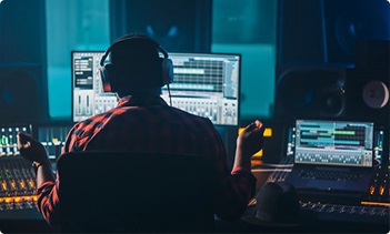 A person wearing headphones sits at a mixing console in a dimly lit recording studio, working with audio editing software displayed on multiple monitors and a laptop.