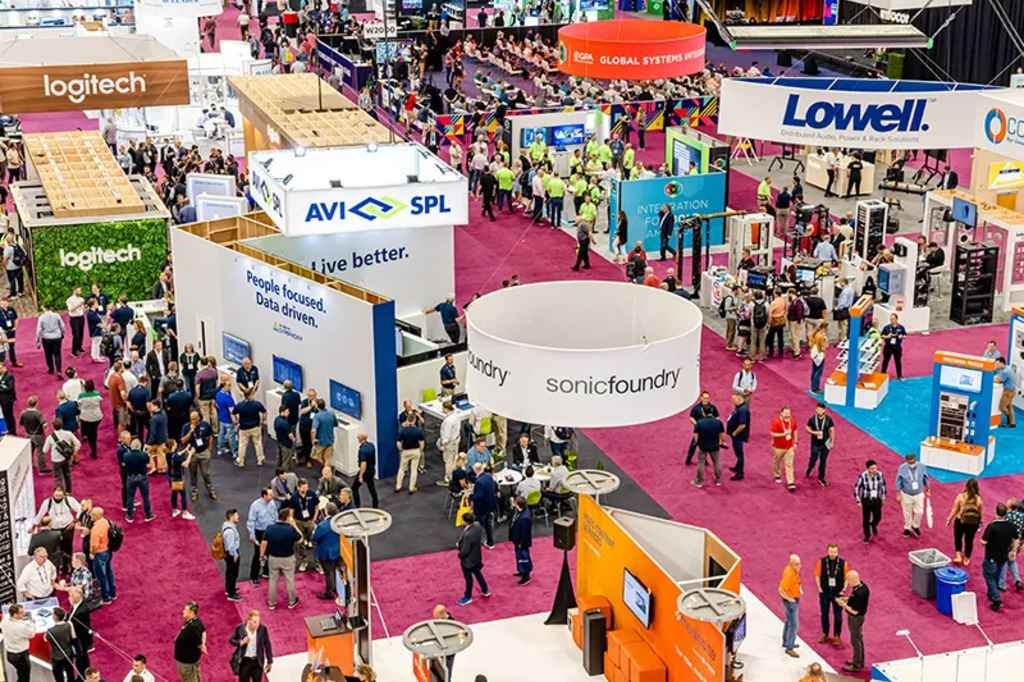 A busy trade show floor with various company booths, colorful signage, and large crowds of people walking, networking, and exploring exhibits. Companies like Logitech, AVI-SPL, and Lowell have visible booths.