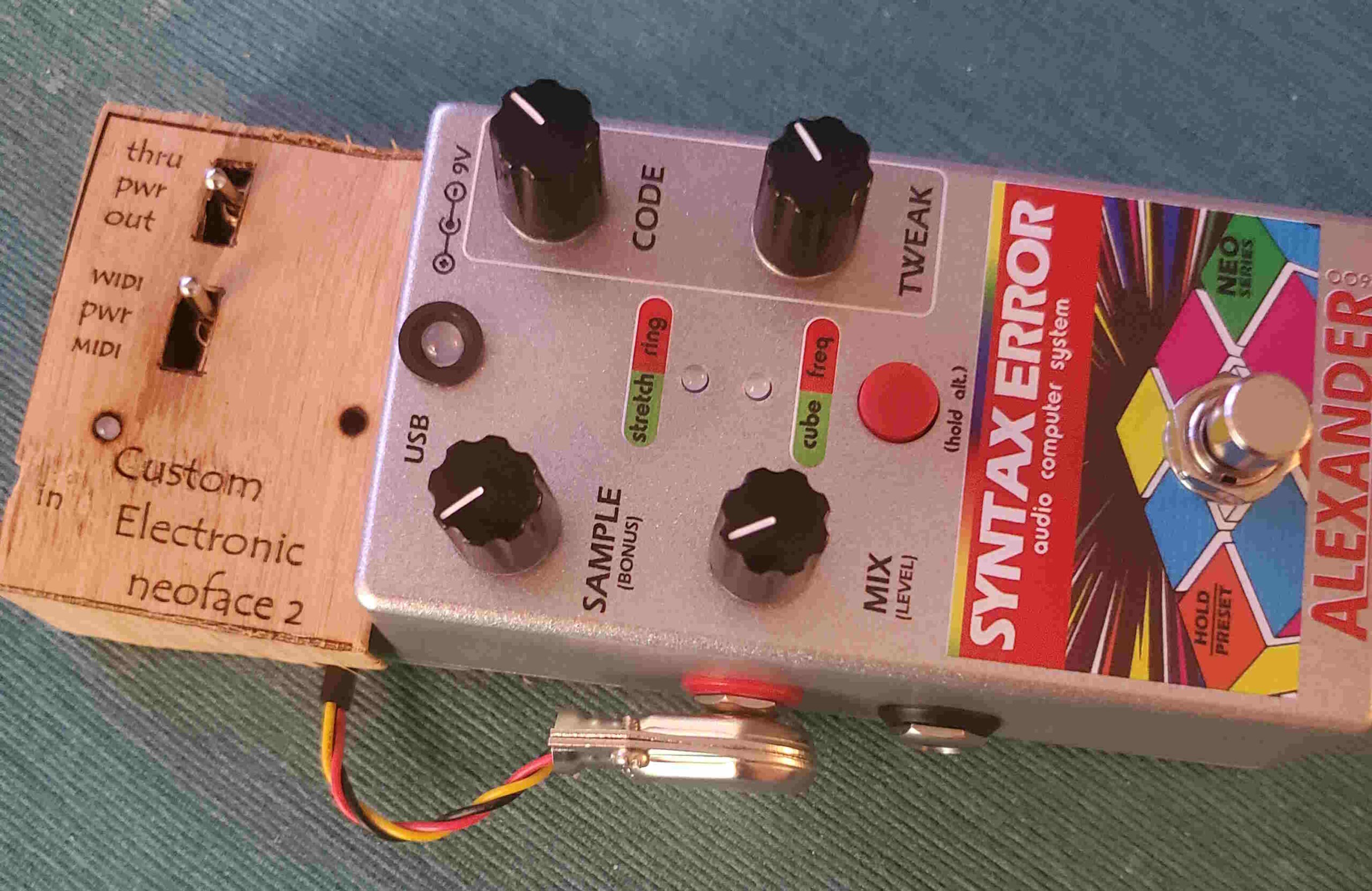 A silver Alexander Syntax Error guitar pedal with colorful labels and knobs is connected to a wooden custom electronic box labeled neoface 2. Cables are attached to both devices, lying on a blue surface.