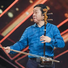 A man in a blue shirt plays a traditional Chinese string instrument called the erhu, holding a bow in one hand, with a blurred stage background featuring diagonal lights.