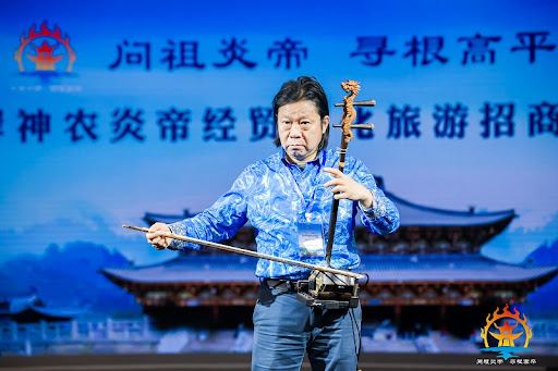A person in a blue shirt plays a traditional string instrument on stage, with Chinese characters and a temple background displayed on a screen behind them. Logos are visible in the bottom corners.