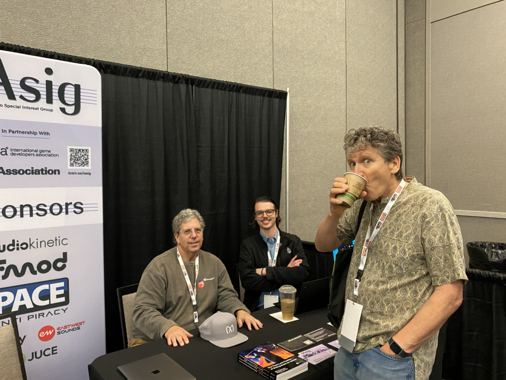 Three men at a conference booth. Two are seated at a table with laptops, drinks, and brochures, smiling at the camera. One stands in front, sipping a drink. A sign with various company logos is visible on the left.