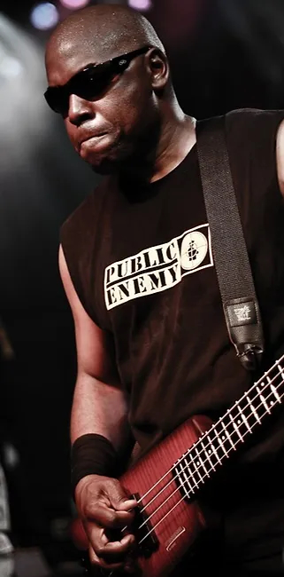 A musician wearing sunglasses and a sleeveless “Public Enemy” shirt plays an electric bass guitar on stage, with dramatic lighting highlighting his focused expression.