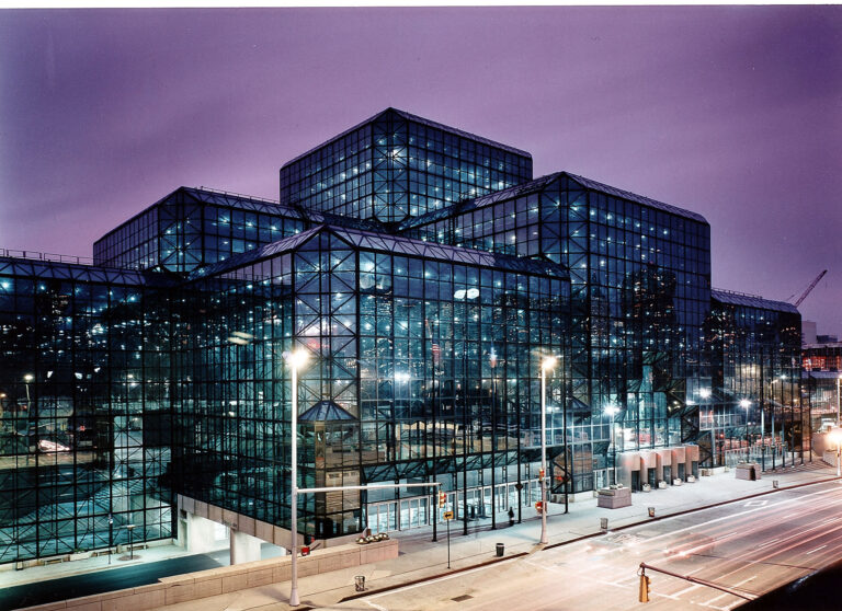A large, modern building with multiple glass and steel sections is lit up at night. The street in front is empty, and the sky has a purple hue. The architecture features geometric shapes and a grid-like glass facade.