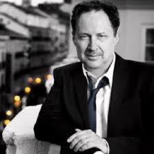 A man in a suit and loosened tie leans casually on a ledge, with a blurred city street and lights in the background. The photo is mostly black and white, with some color in the background lights.