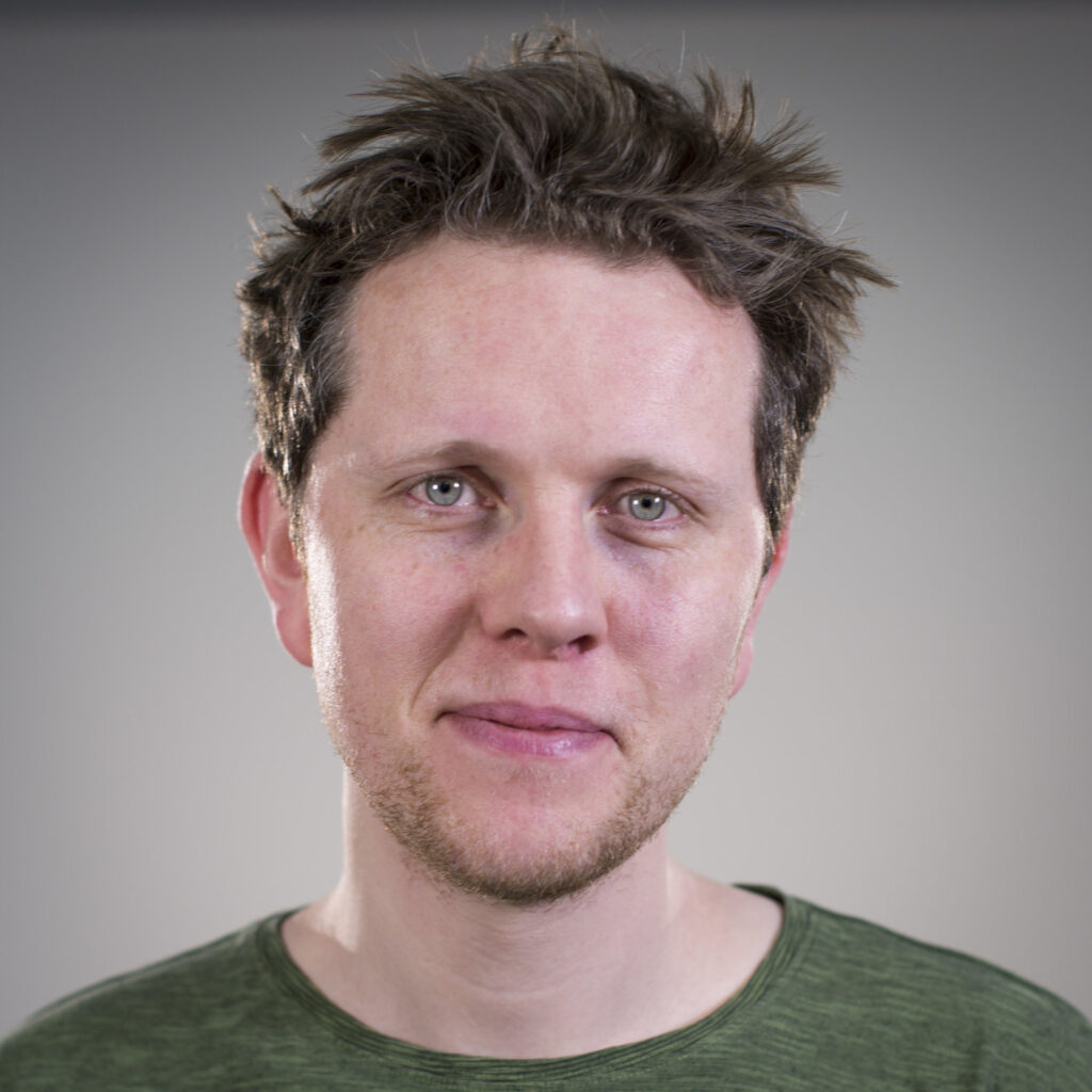 A man with short, tousled brown hair and light skin is wearing a green shirt, looking directly at the camera with a neutral expression. The background is plain and softly lit.