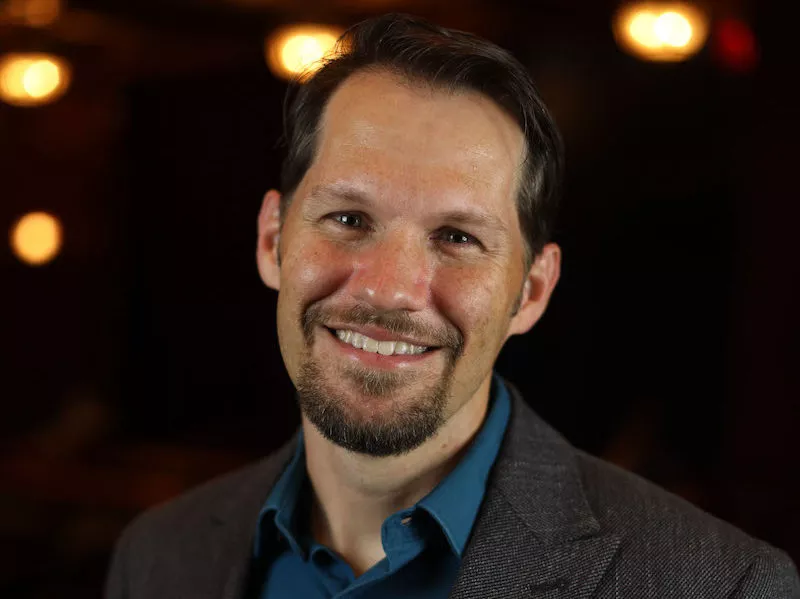 A man with short dark hair and a trimmed beard smiles at the camera, wearing a blue shirt and gray blazer. Warm, soft lights glow in the blurred background.
