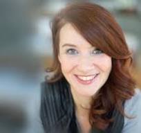 A woman with long, wavy auburn hair and light skin is smiling at the camera. She is wearing a dark top and is photographed in soft-focus lighting with a blurred background.