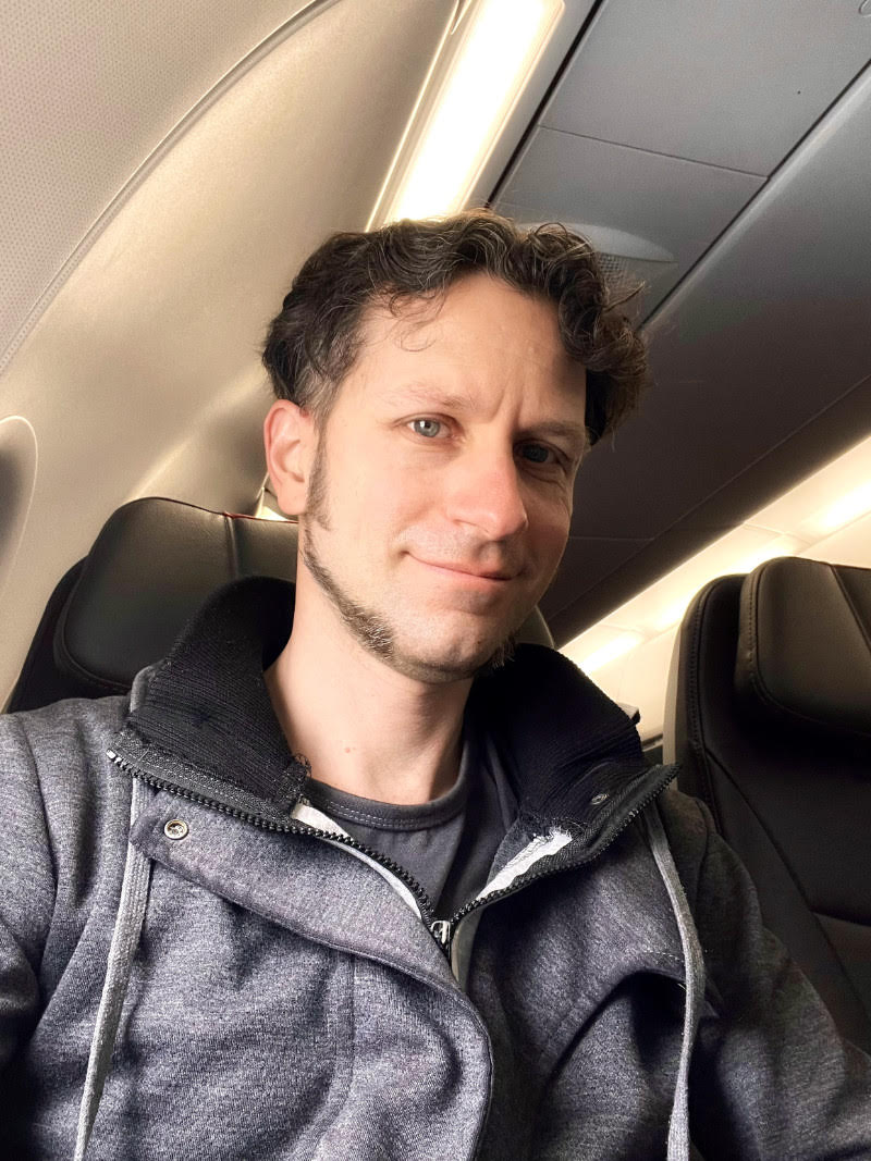 A man with short, curly brown hair and light eyes smiles while seated on an airplane. He is wearing a gray zip-up jacket and a dark shirt. The background shows a window and empty seat beside him.