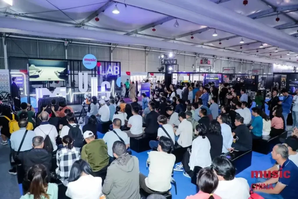 A large audience sits and watches a presentation or performance on stage in a spacious, brightly lit indoor venue at Music China. Various booths and displays are visible in the background.