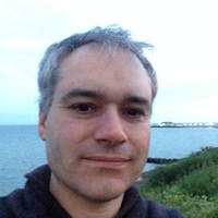 A person with short gray hair is smiling slightly while standing outdoors near water, with grass and the horizon visible in the background under a pale sky.