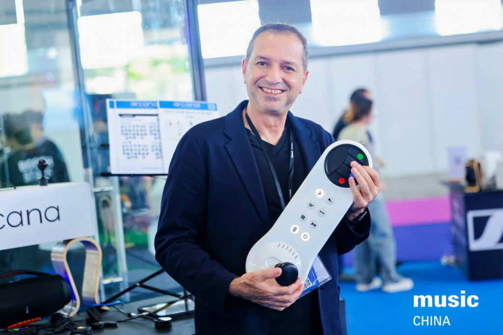 A smiling man in a black blazer holds a large, white electronic device with buttons and dials at a trade show. There are blurred people and booths in the background, with music CHINA text in the corner.