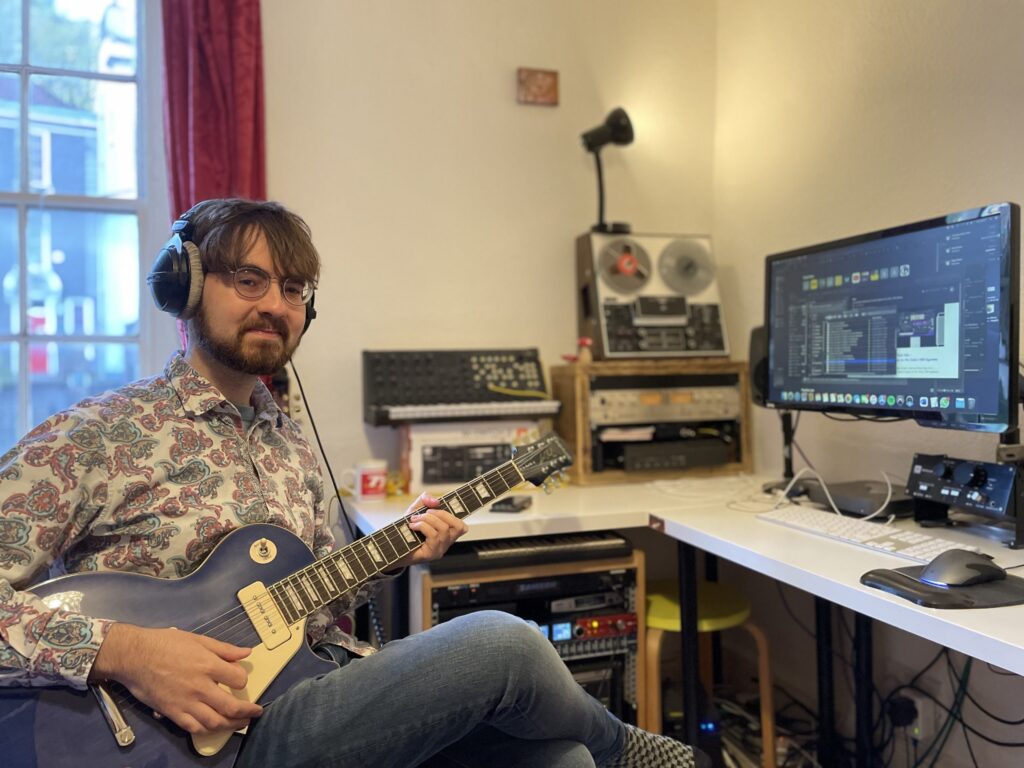 A man wearing headphones and a patterned shirt plays an electric guitar in a home studio, surrounded by audio equipment and a computer displaying music production software.