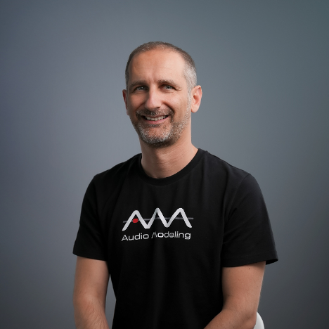 A smiling man with short hair and a beard sits on a stool against a plain gray background, wearing a black Audio Modeling t-shirt with a white and red logo.