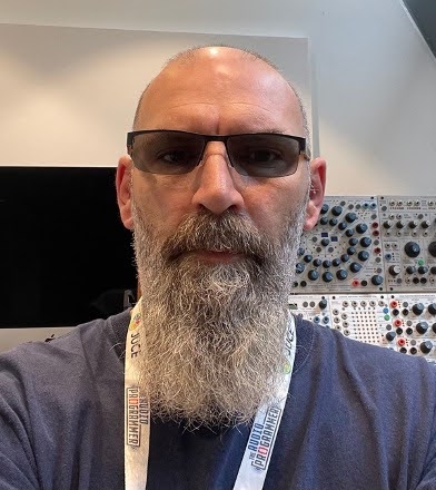 A man with a long gray beard and sunglasses is wearing a lanyard and a blue shirt, standing indoors in front of a wall of modular synthesizer equipment.