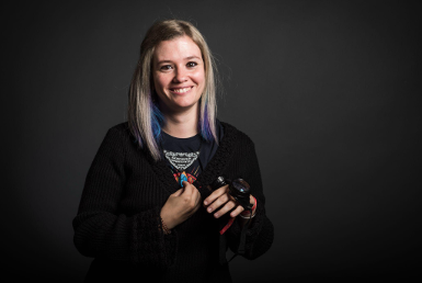 A smiling woman with light hair and purple streaks stands against a dark background, wearing a black sweater and holding a camera in one hand and a small toy or trinket in the other.