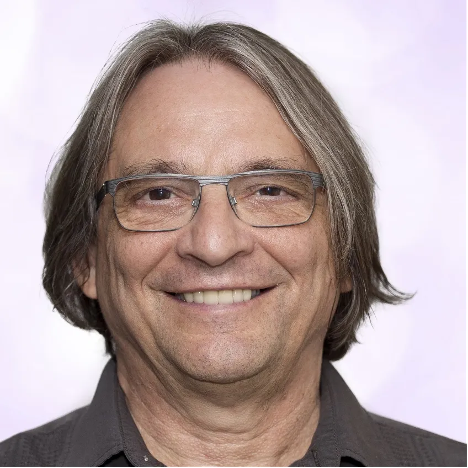 A middle-aged man with straight, shoulder-length gray hair and glasses smiles at the camera. He is wearing a dark collared shirt and the background is a soft, light purple.