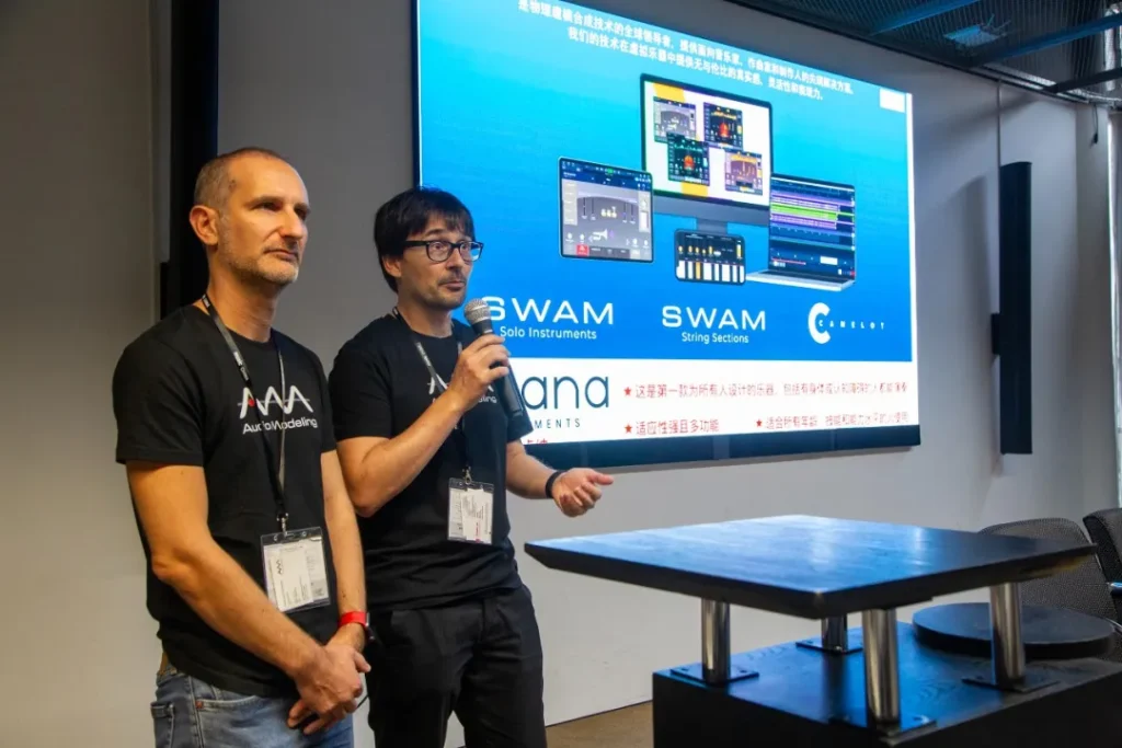 Two men stand in front of a large screen giving a presentation about SWAM Solo Instruments and SWAM String Sections. One man speaks into a microphone while the other listens attentively.