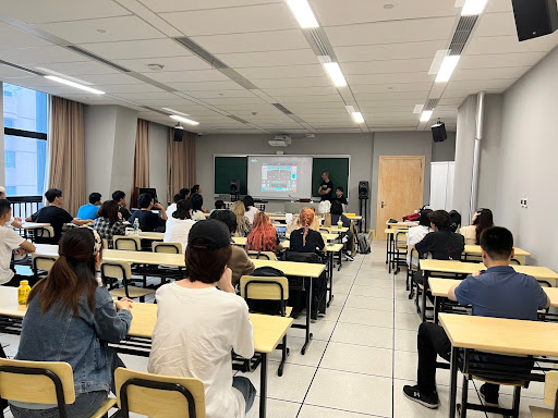 A classroom filled with students sitting at desks facing a teacher who is presenting information on a screen at the front of the room. The room is well-lit with windows on one side.