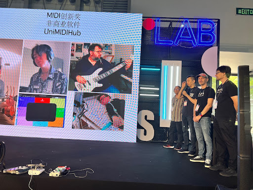 A group of five people stands on a stage next to a large screen displaying photos of individuals with musical equipment and digital graphics, under a sign that says LAB. Electronic devices are visible on the stage floor.