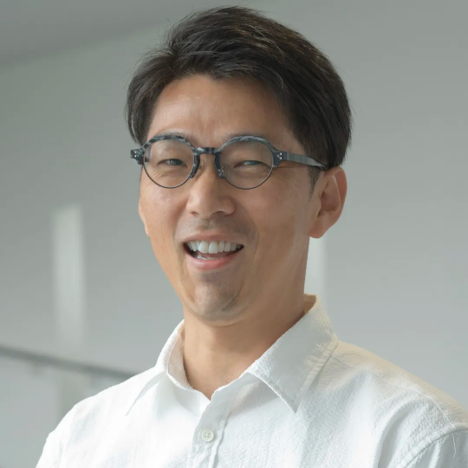 A man with short dark hair and round glasses smiles while wearing a white shirt, standing against a light, blurred background.